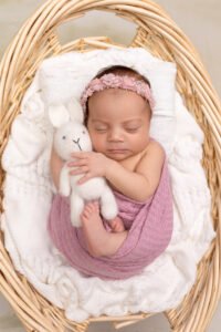 Sleeping newborn baby girl in a pink wrap cradling a knitted bunny toy in a wicker basket, demonstrating the results of a perfectly timed session booked during the second trimester with Tender Beginnings Photo.