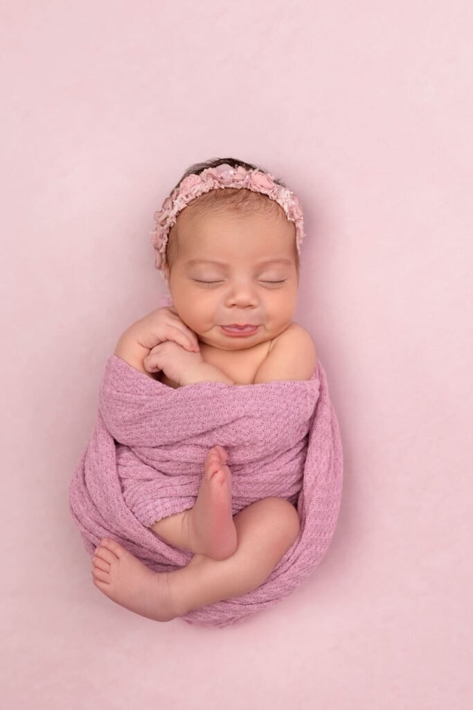 Smiling newborn baby girl wrapped in a soft textured pink swaddle with a matching floral headband at Tender Beginnings studio.