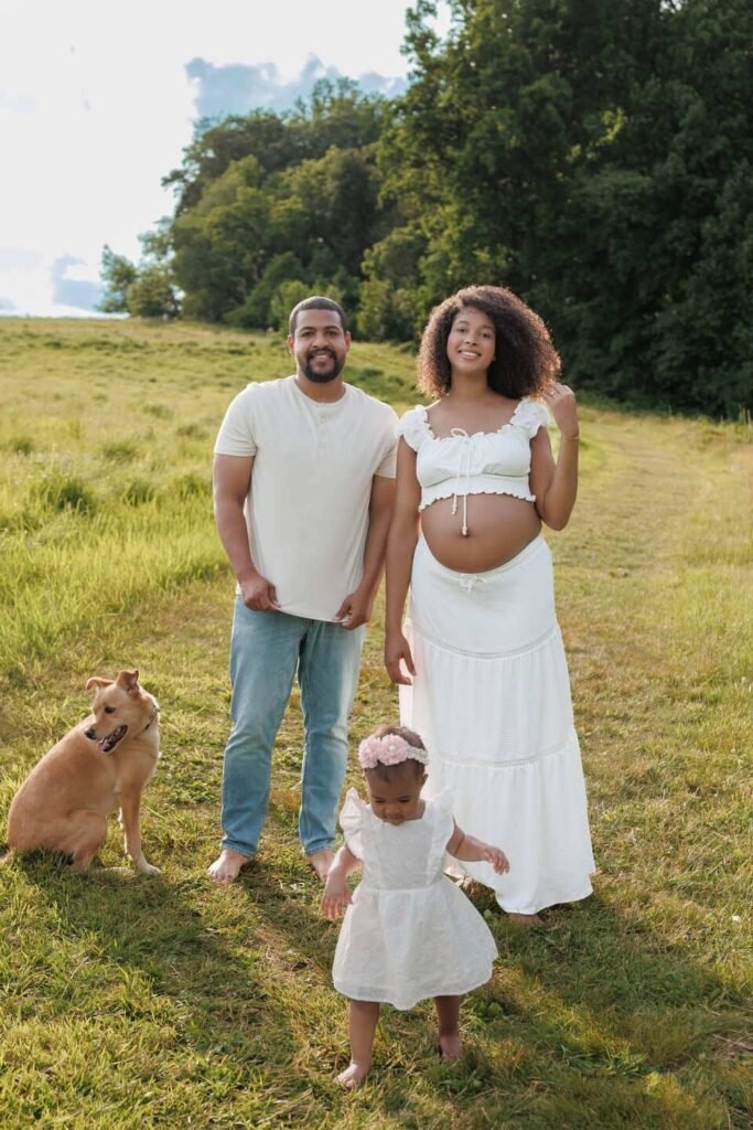Outdoor maternity and family photography session in a scenic field, featuring expecting parents with their toddler and dog in coordinated white outfits, showcasing the airy style of Tender Beginnings Photo.
