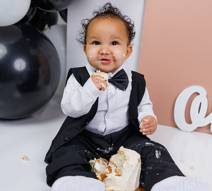 Joyful one-year-old boy in a tuxedo outfit digging into his first birthday cake during a fun, messy cake smash session at our Richboro studio.
