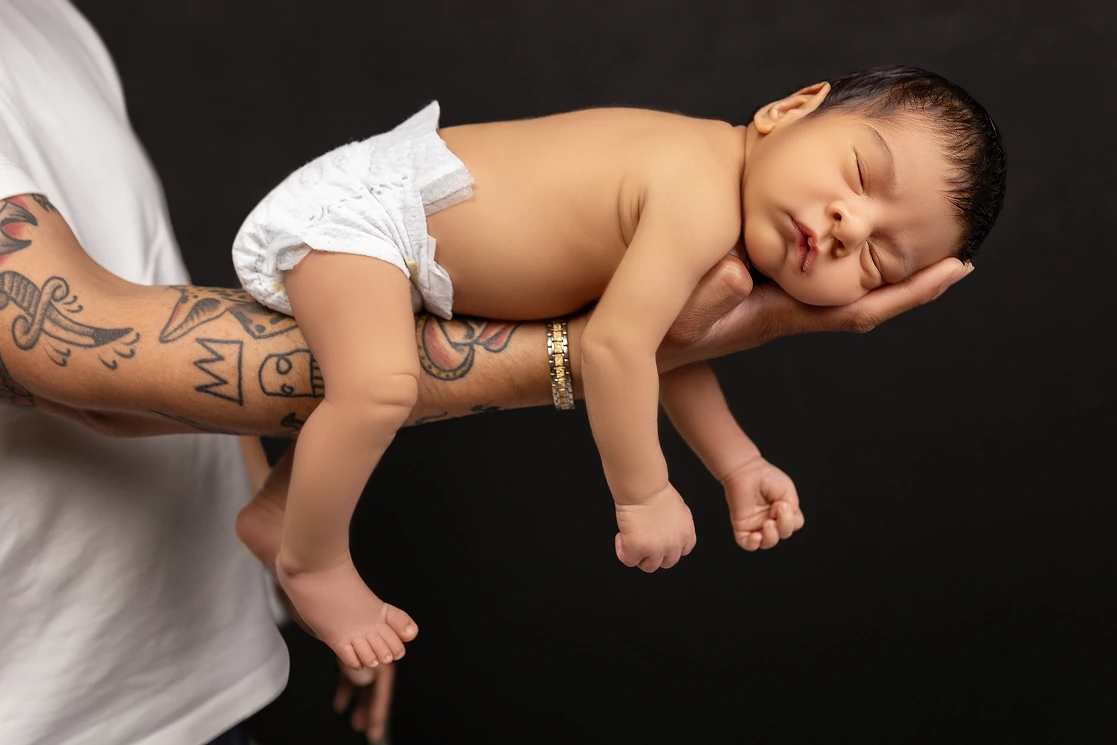 Newborn baby sleeping on father's tattooed arm, studio portrait by Tender Beginning