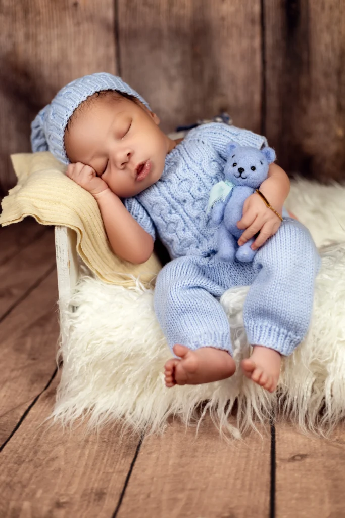 Sleeping newborn boy in a cozy blue knitted outfit holding a tiny bear, captured during a relaxed studio session.