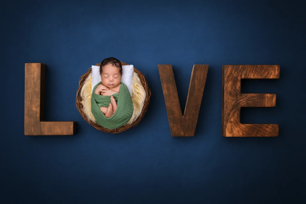 Baby safely swaddled in green and posed inside a rustic wooden LOVE sign against a dark blue background.