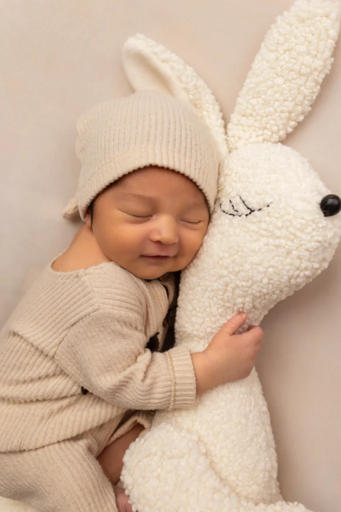 Newborn baby in a neutral beige outfit comfortably hugging a large plush bunny during a baby-led session in PA.