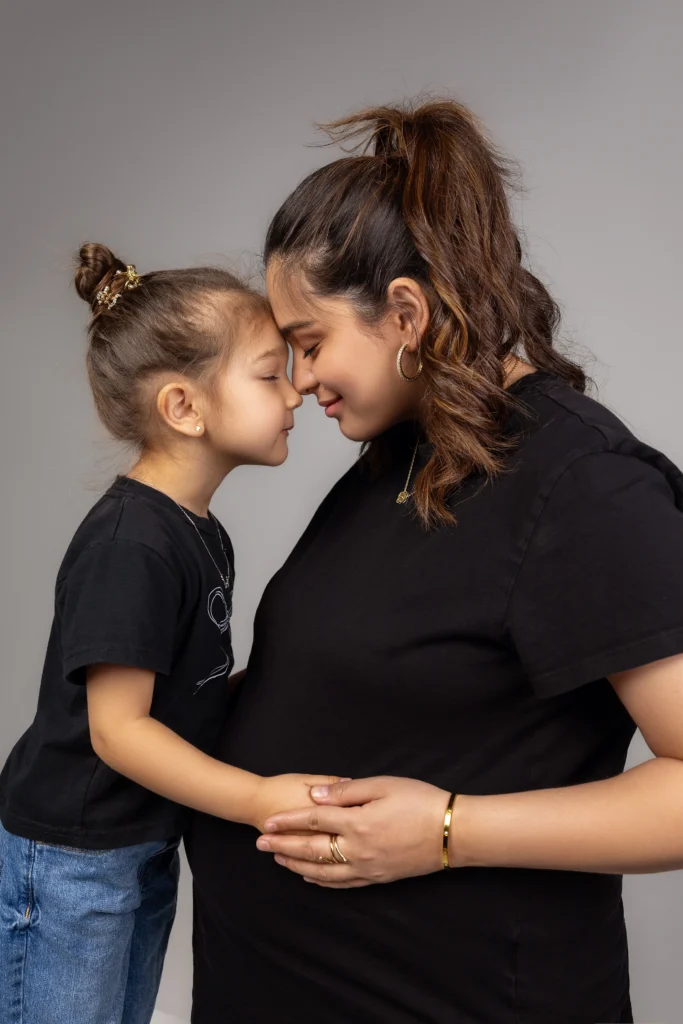 Mother and young daughter sharing a sweet forehead-to-forehead moment while touching the baby bump in casual black tees.