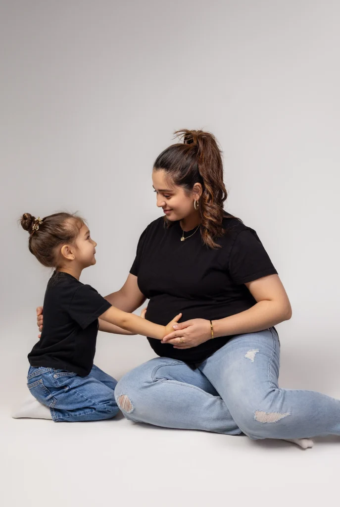 Relaxed mother and daughter sitting cross-legged on the studio floor sharing giggles while touching the baby bump.