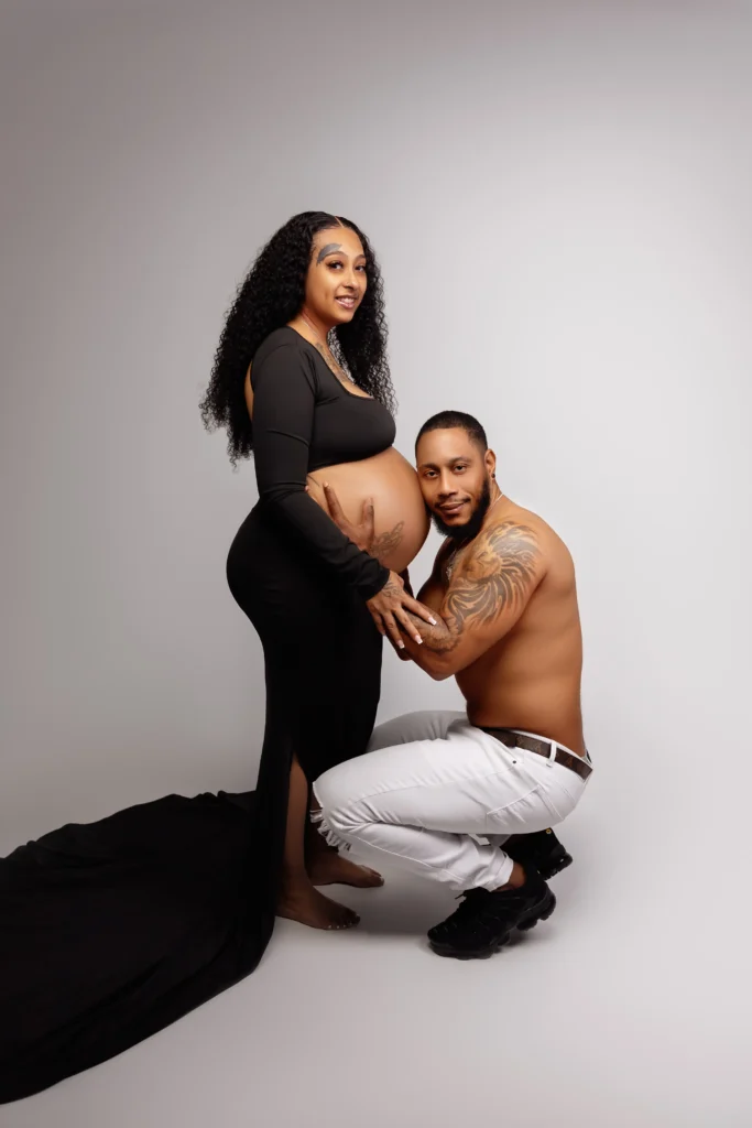 Expecting mother in a sleek black dress with her husband kneeling beside her to embrace the baby bump in our studio.