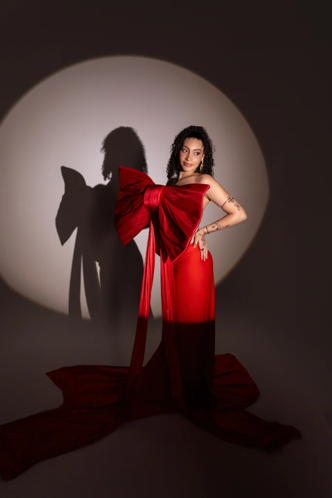 Dramatic editorial maternity portrait featuring a bold red gown with an oversized bow illuminated by a studio spotlight.