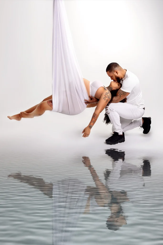 Creative studio maternity photo of an expecting mother suspended in white silks with water-like floor reflection and her husband.
