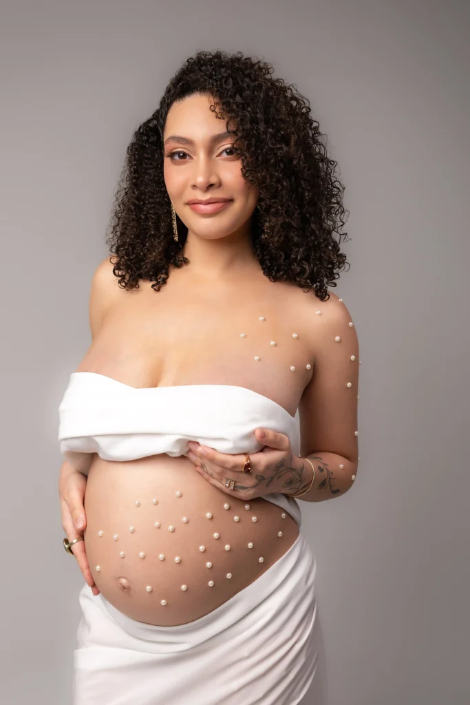 Glowing pregnant mother draped in minimal white fabric with delicate adhesive pearls accenting her shoulders and bump.