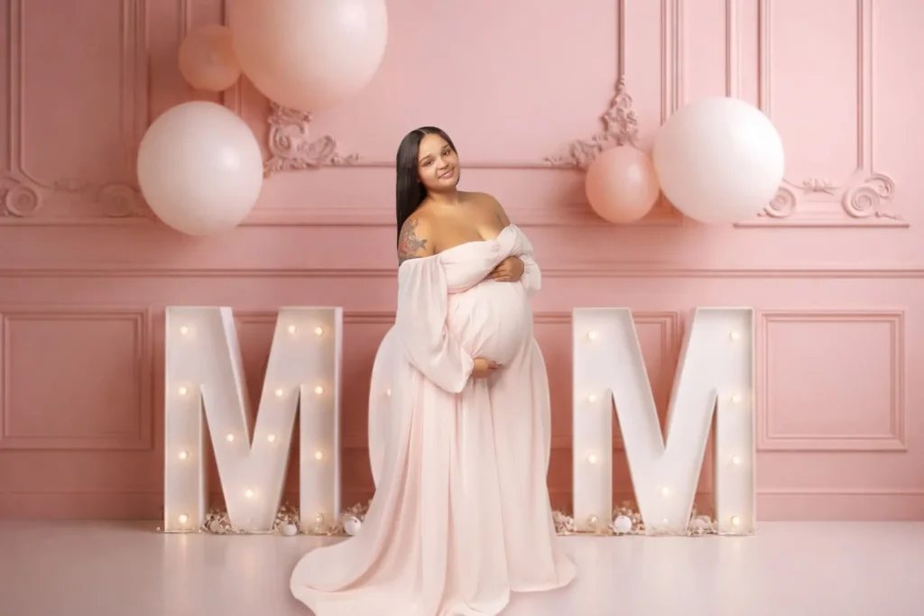 Pregnant woman in an off-the-shoulder pink gown standing in front of illuminated marquee MOM letters and pastel balloons.