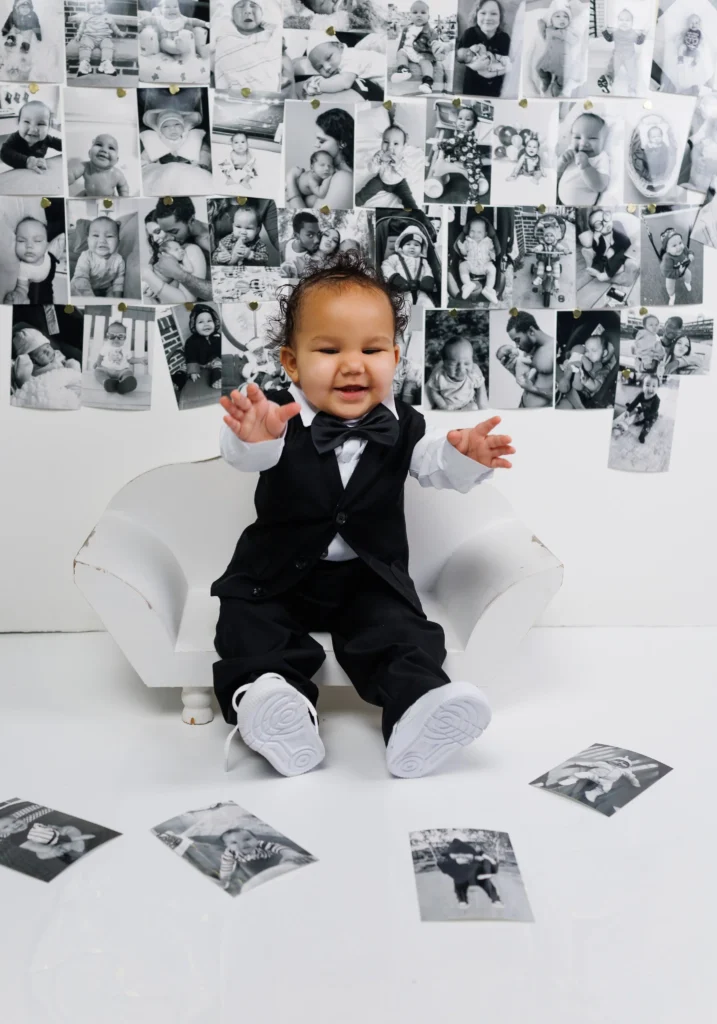 One-year-old boy in a black tuxedo sitting in front of a black-and-white milestone photo collage celebrating his first year.