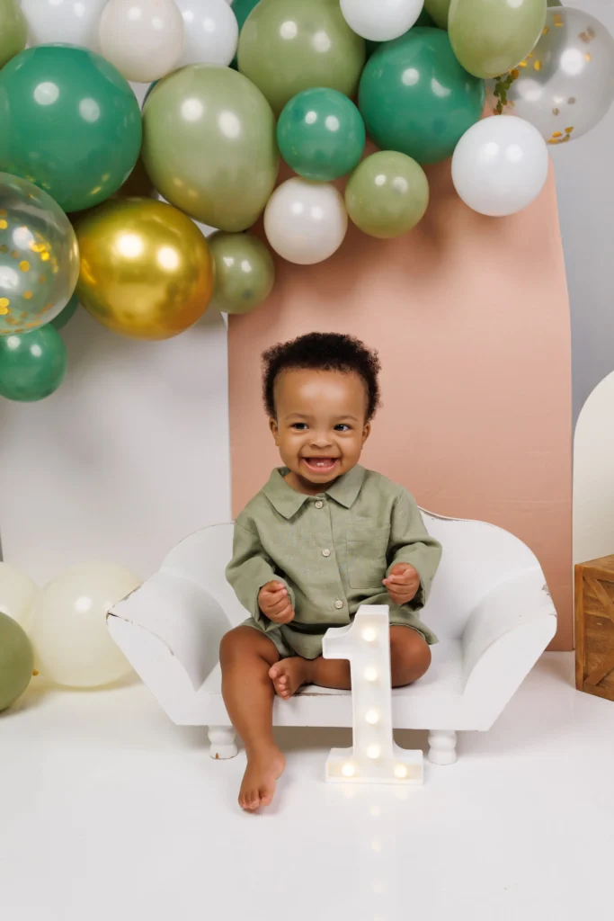 Smiling baby boy in a sage green romper sitting next to a light-up marquee number one with olive and gold balloon decor in our studio.