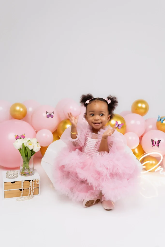 Happy baby girl with pigtails wearing a fluffy pink tulle dress surrounded by pastel pink and gold balloons for her first birthday.
