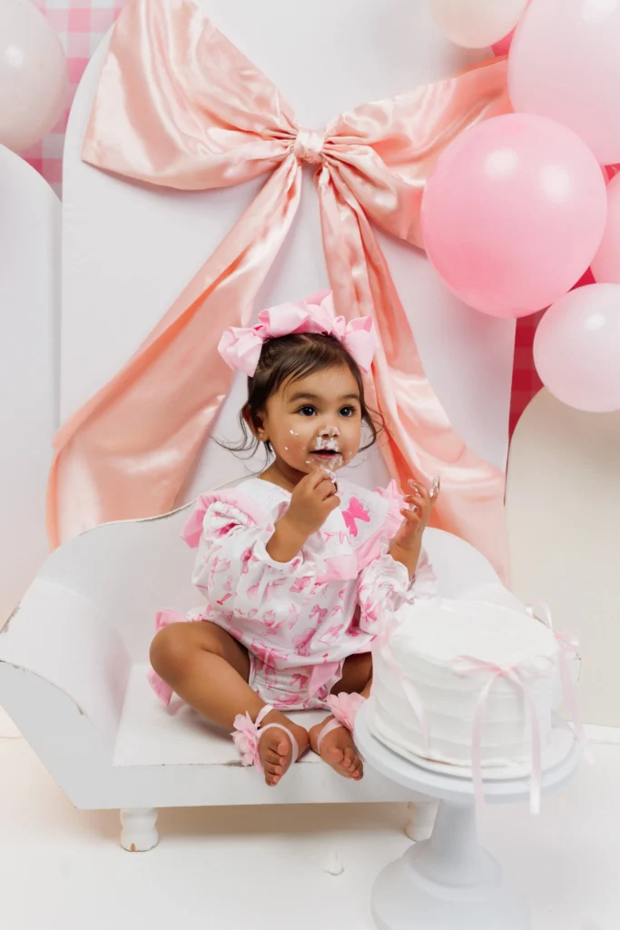 Sweet one-year-old girl tasting white cake frosting in front of an oversized peach bow backdrop during a Philadelphia milestone session.