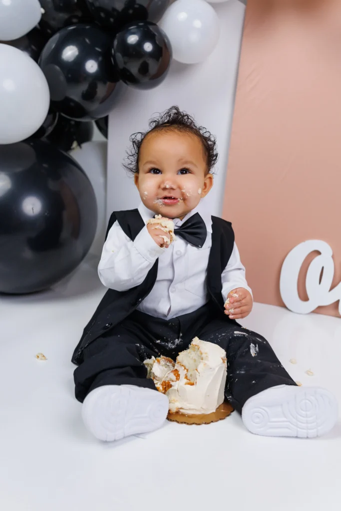 Joyful baby boy in a black bow tie tasting vanilla frosting with messy hands, fully embracing the fun of his first birthday.