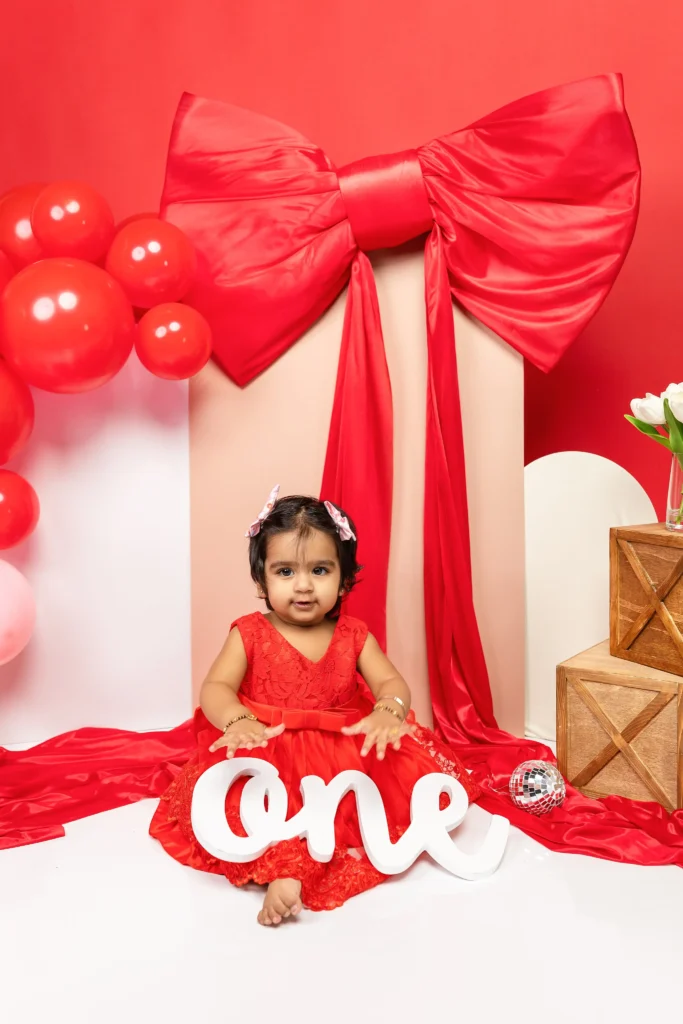 Little girl in a vibrant red dress seated in front of a giant red bow and white wooden "one" sign, proving we love bold birthday colors.