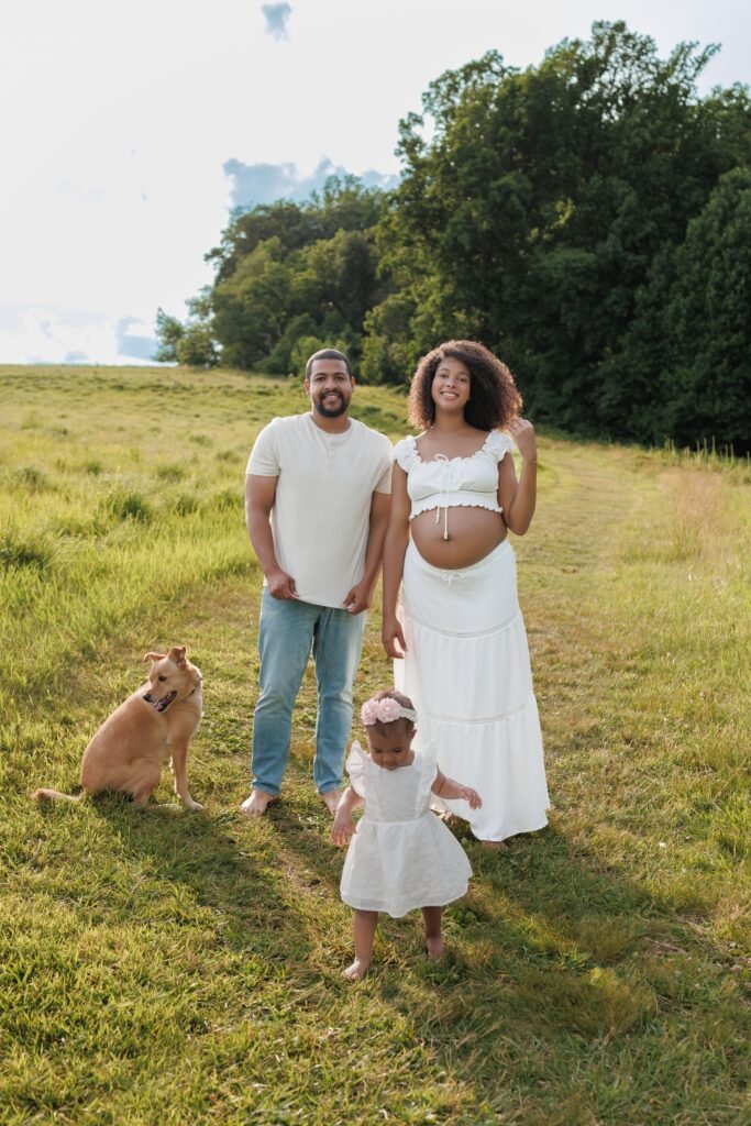 Outdoor maternity and family photography session in a scenic field, featuring expecting parents with their toddler and dog in coordinated white outfits, showcasing the airy style of Tender Beginnings Photo.