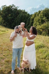 Candid outdoor maternity photography moment with a father holding a toddler on his shoulders next to his pregnant wife, illustrating the natural lifestyle feel of a sunset field session.