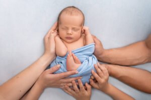 Sleeping newborn baby boy in a light blue wrap surrounded by family hands during a professional studio session at Tender Beginnings Photo, showcasing luxury newborn photography styling.