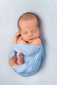 Serene newborn baby boy wrapped in soft blue fabric, professionally posed to ensure safety and comfort in a temperature-controlled studio environment.