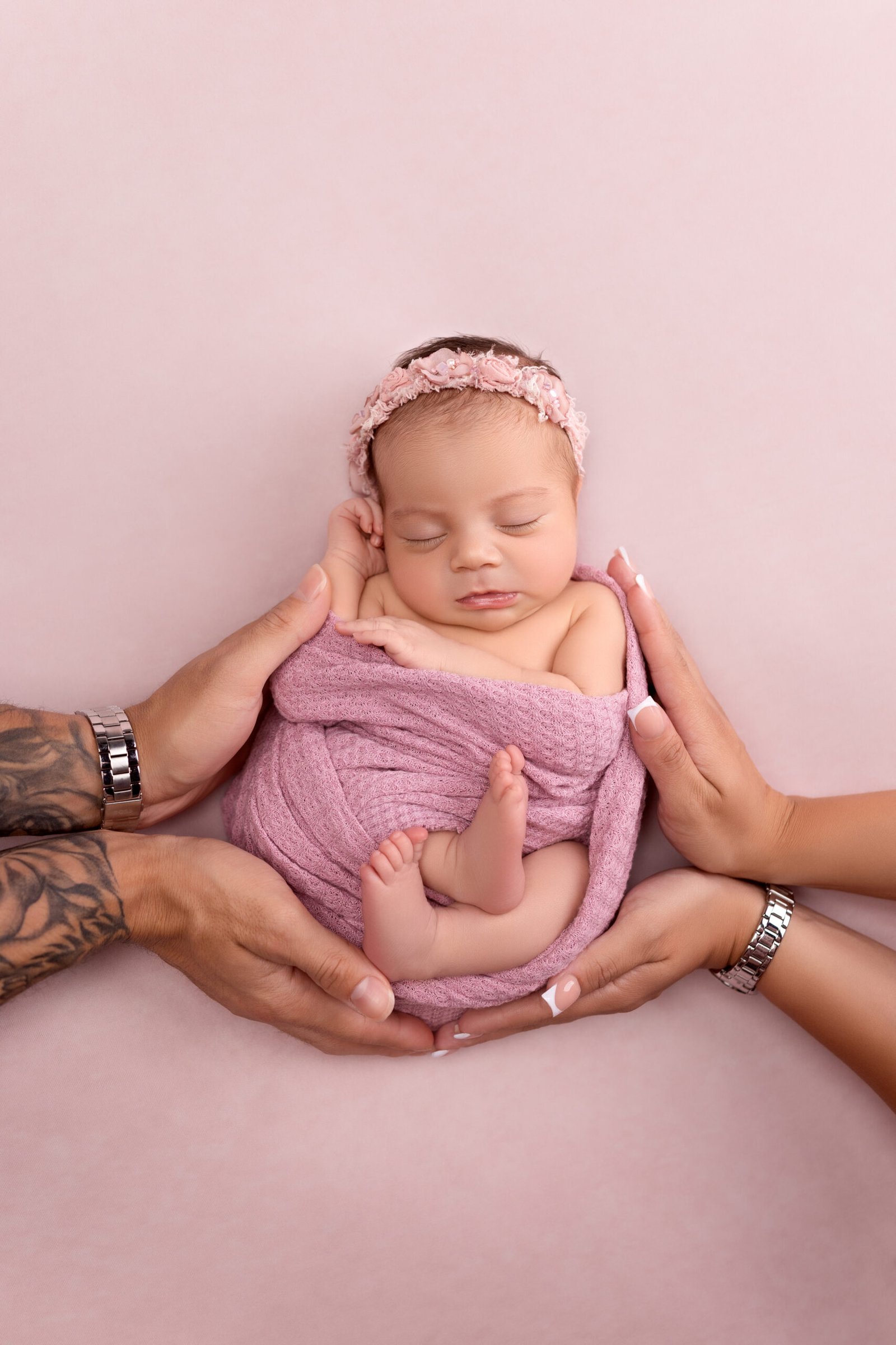Sleeping newborn baby girl in a pink wrap safely cradled in her parents' hands during a professional studio session at Tender Beginnings Photo.