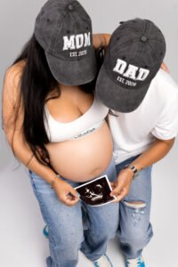 Modern studio maternity photography featuring a couple in casual denim and custom "Mom and Dad" hats holding an ultrasound photo, highlighting the clean and personalized aesthetic of indoor sessions.