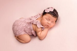 Sleeping newborn baby girl in a luxury hand-knitted pink romper and matching headband, showcasing the boutique studio wardrobe available at Tender Beginnings Photo.